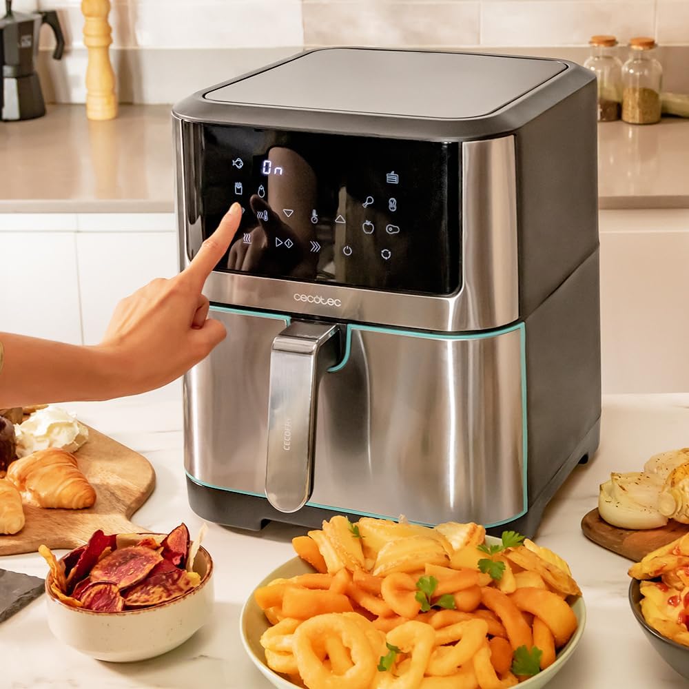 Doigt qui effleure le bandeau tactile du Cecotec Cecofry Supreme 8000 en inox, entouré de frites, chips de betterave et croissants dorés sur un plan de travail clair