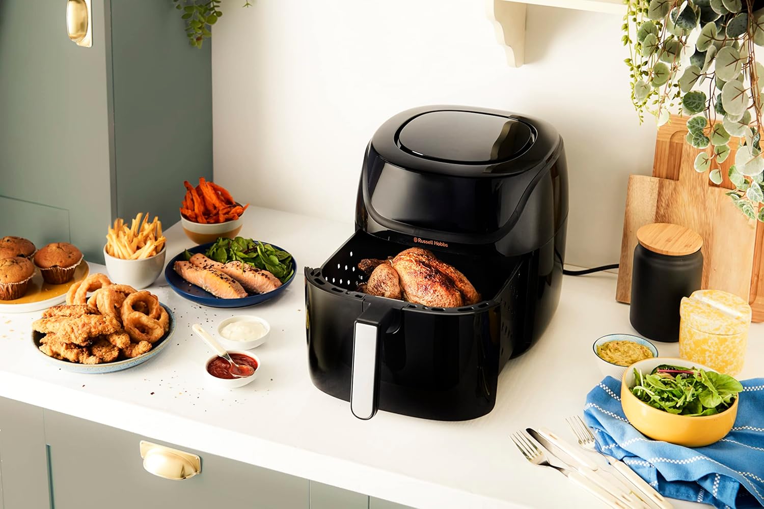 Panier du Russell Hobbs SatisFry 27160-56 tiré hors de l'appareil avec un poulet rôti à l'intérieur, entouré d'assiettes de frites, de saumon et de muffins