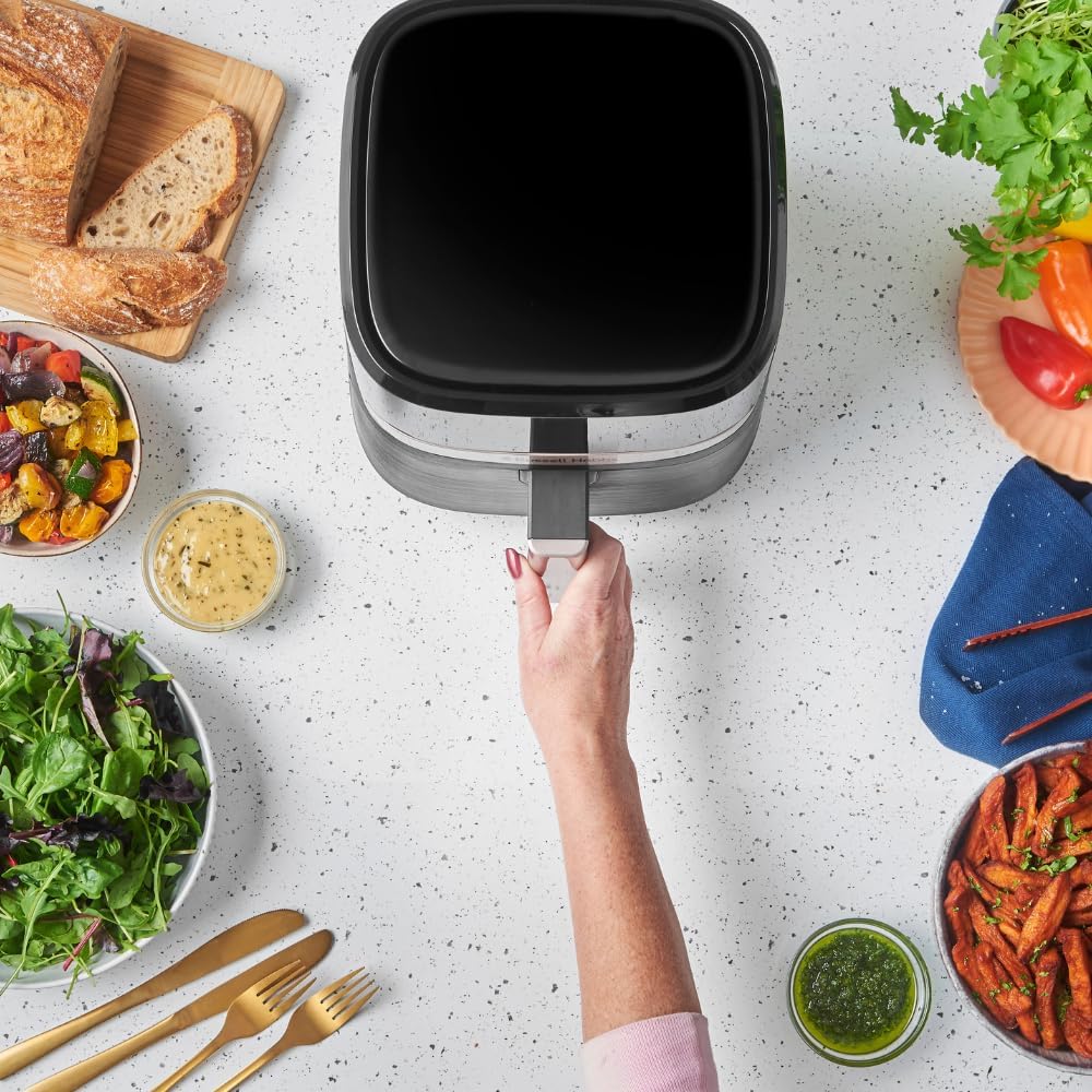 Vue de dessus du Russell Hobbs SatisFry 27610-56 posé sur un plan de travail moucheté, une main tirant la poignée du panier au milieu de plats servis