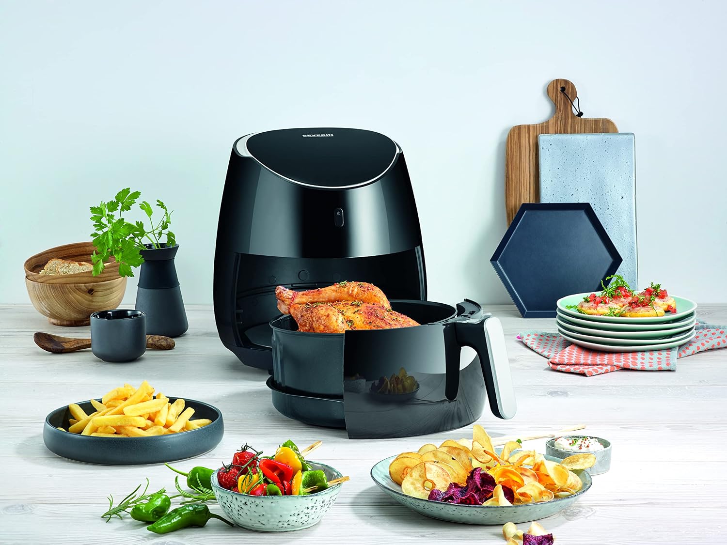 Airfryer Severin FR 2445 posé sur un plan de travail clair, panier sorti sur un poulet entier doré, entouré d'assiettes de frites, de chips de légumes et de brochettes