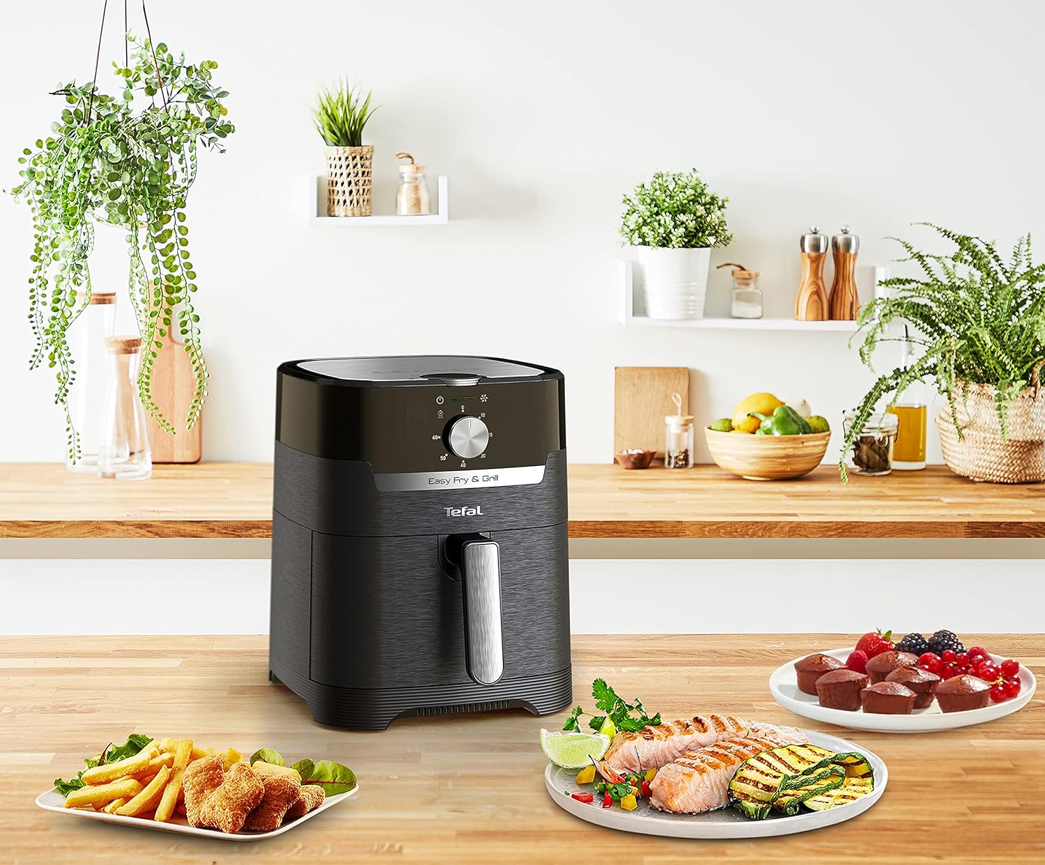 Tefal EY5018 Easy Fry & Grill noir sur un plan de travail, entouré d'assiettes de saumon et courgettes grillés, de frites et de muffins
